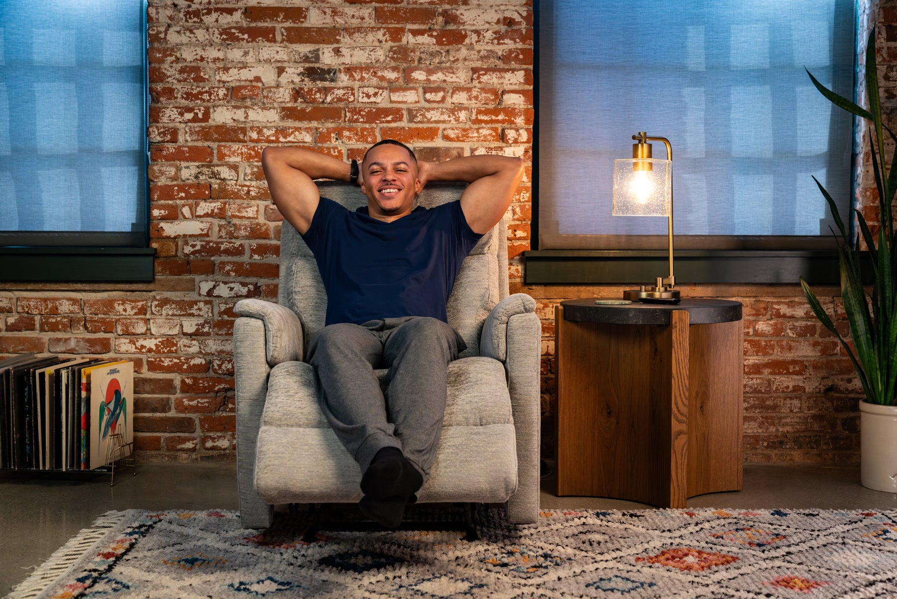 Man relaxing in a light gray recliner chair with arms behind his head, smiling in a cozy living room with brick wall, side table and lamp.”